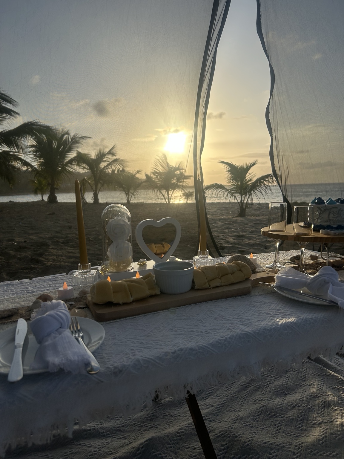 Anniversary picnic table beneath a white canopy beside the beach at sunset, with candles, flowers and place settings